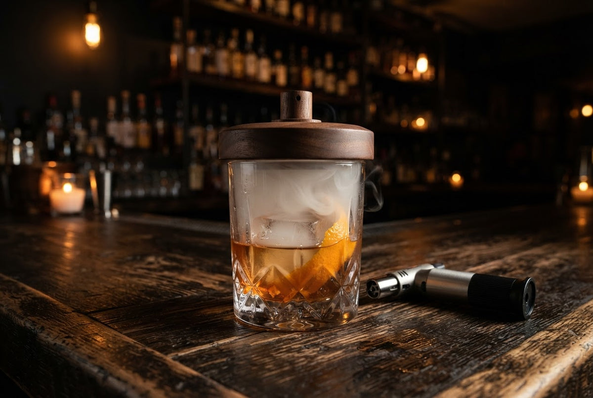 A smoked Old Fashioned with an orange peel and a large ice cube under a wooden cocktail smoker, with a torch beside it
